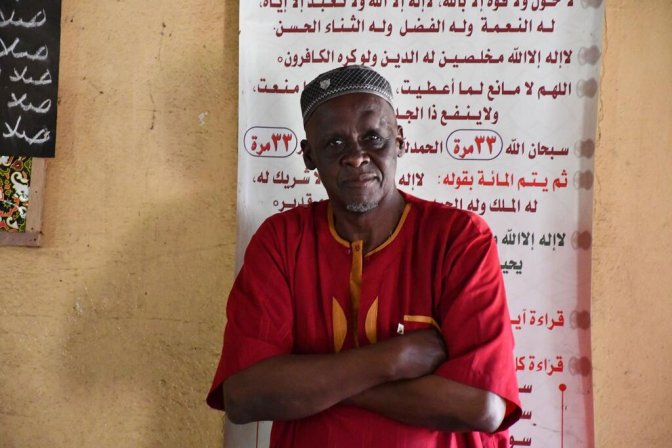 Un hombre con una camisa roja y una tapa de cabeza se encuentra con los brazos cruzados frente a una pared con texto árabe, simbolizando la resistencia de la comunidad contra la mutilación genital femenina en Guinea.