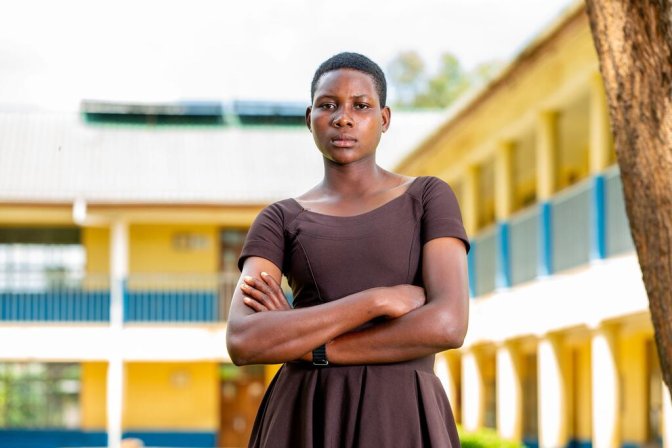 Una joven está de pie con confianza con los brazos cruzados frente a un edificio de la escuela, simbolizando la resistencia y la resistencia contra la mutilación genital femenina en Tanzania.